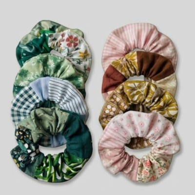 Duas colunas de elásticos de cabelo (scrunchies) feitos à mão, dispostos verticalmente sobre um fundo cinzento claro. A coluna da esquerda apresenta tons de verde e azul com padrões botânicos, xadrez e riscas. A coluna da direita exibe tons de rosa, castanho e mostarda com estampados florais e retro. Os tecidos têm texturas variadas, sugerindo um estilo artesanal e vintage