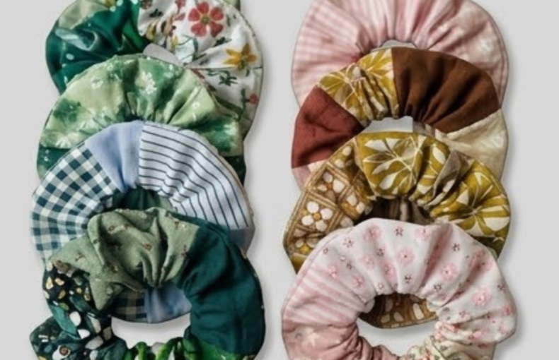 Duas colunas de elásticos de cabelo (scrunchies) feitos à mão, dispostos verticalmente sobre um fundo cinzento claro. A coluna da esquerda apresenta tons de verde e azul com padrões botânicos, xadrez e riscas. A coluna da direita exibe tons de rosa, castanho e mostarda com estampados florais e retro. Os tecidos têm texturas variadas, sugerindo um estilo artesanal e vintage