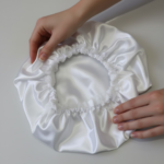 Uma touca de cetim branca brilhante pousada sobre uma superfície clara, sendo segurada por duas mãos femininas que demonstram a elasticidade e a textura suave do tecido.