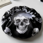 Vista superior de uma touca de dormir em cetim preto com padrão repetido de caveiras brancas detalhadas. O material é brilhante e tem um elástico franzido na abertura.