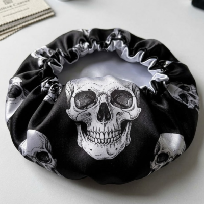 Vista superior de uma touca de dormir em cetim preto com padrão repetido de caveiras brancas detalhadas. O material é brilhante e tem um elástico franzido na abertura.