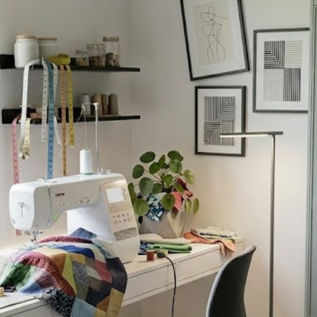 Máquina de costura branca sobre uma secretária moderna, com um trabalho de "patchwork" colorido a ser costurado. Ao fundo, prateleiras com frascos e fitas métricas penduradas, uma planta de interior e quadros com arte minimalista na parede branca.
