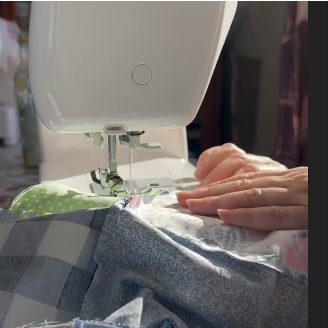 Uma fotografia em grande plano que mostra as mãos de uma pessoa a guiar cuidadosamente tecidos com padrões (xadrez azul e flores) sob a agulha de uma máquina de costura branca e moderna. A luz natural ilumina a área de trabalho.