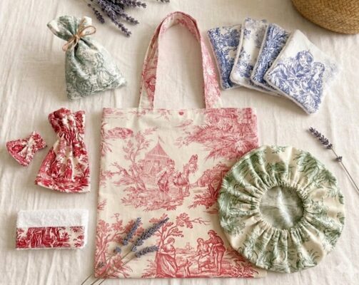 Uma foto aérea de estúdio, em um fundo de linho texturizado, exibe uma coleção de itens de Toile de Jouy em cores variadas. No centro está uma tote bag grande com padrão Toile de Jouy vermelho e off-white. Ao redor dela, estão dispostos: um sachê de perfume verde, um pequeno laço vermelho, uma bolsa de cordão vermelha, um rolo de toalha de mão vermelho, uma touca de banho elástica verde, um conjunto de cinco ecopads azuis e alguns ramos de lavanda seca.