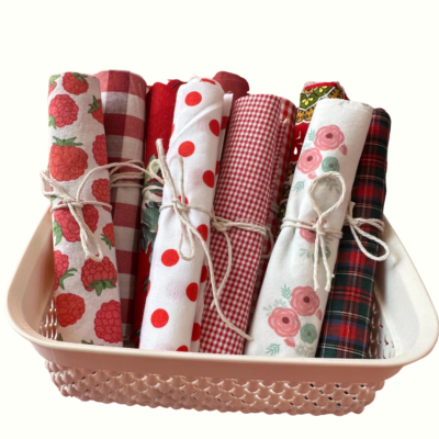 Lote de rolos de tecido de algodão em tons de vermelho, branco e padrões florais, atados com cordel rústico e dispostos dentro de um cesto branco perfurado.
