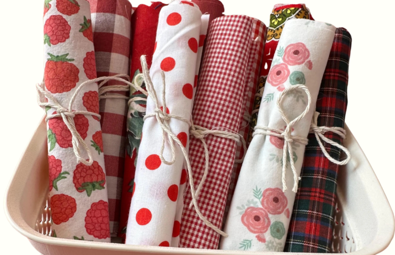 Lote de rolos de tecido de algodão em tons de vermelho, branco e padrões florais, atados com cordel rústico e dispostos dentro de um cesto branco perfurado.
