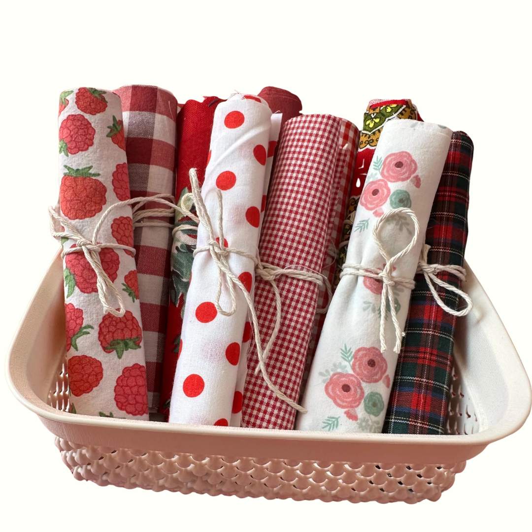Lote de rolos de tecido de algodão em tons de vermelho, branco e padrões florais, atados com cordel rústico e dispostos dentro de um cesto branco perfurado.