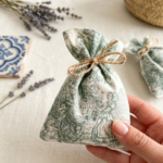 "Uma mão segura um pequeno saco de alfazema artesanal com padrão Toile de Jouy em verde. Ao fundo, em cima de uma mesa de linho claro, vêem-se ramos de alfazema seca, um azulejo tradicional português azul e branco e um cesto de verga, criando um ambiente rústico e acolhedor."