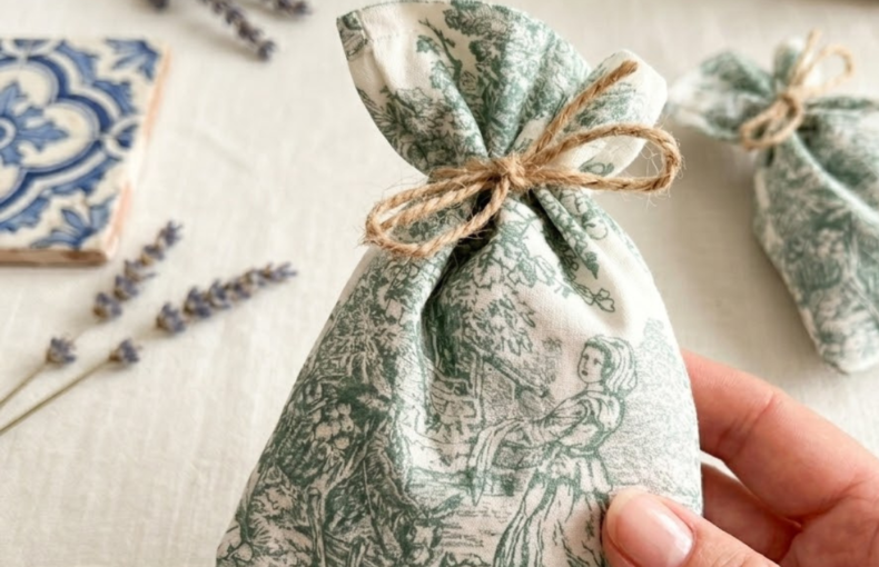 "Uma mão segura um pequeno saco de alfazema artesanal com padrão Toile de Jouy em verde. Ao fundo, em cima de uma mesa de linho claro, vêem-se ramos de alfazema seca, um azulejo tradicional português azul e branco e um cesto de verga, criando um ambiente rústico e acolhedor."
