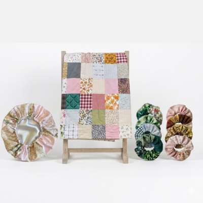 Uma colcha de retalhos colorida estendida num suporte de madeira, ladeada por uma touca de cetim floral e duas pilhas de elásticos de cabelo (scrunchies) em tons de verde, rosa e castanho, tudo sobre um fundo branco minimalista.
