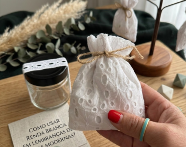 Uma mão feminina com unhas vermelhas e um anel turquesa segura uma pequena bolsa de renda branca amarrada com barbante de juta. No fundo, há um frasco de vidro com tampa preta, um cartão com texto e um selo de cera verde e outras bolsas penduradas em um suporte de madeira. A cena é em uma mesa de madeira com pampas secos.