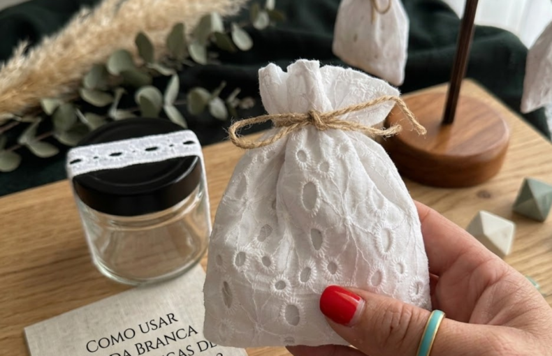Uma mão feminina com unhas vermelhas e um anel turquesa segura uma pequena bolsa de renda branca amarrada com barbante de juta. No fundo, há um frasco de vidro com tampa preta, um cartão com texto e um selo de cera verde e outras bolsas penduradas em um suporte de madeira. A cena é em uma mesa de madeira com pampas secos.