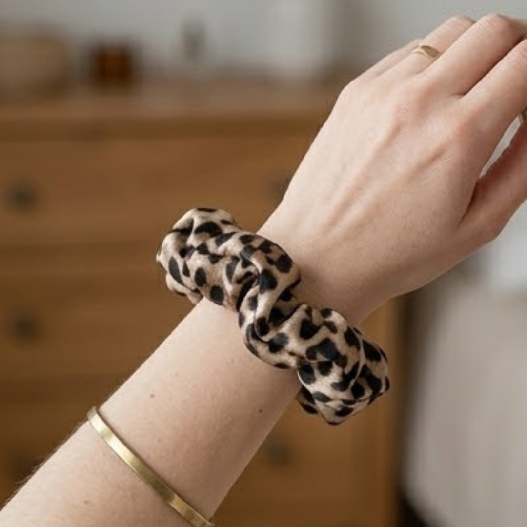scrunchie-cetim-leopardo-artesanal-clotilde-studio.jpg No pulso: Close-up de um scrunchie de cetim com padrão de leopardo usado no pulso como pulseira, junto a uma pulseira dourada fina. No cabelo: Vista posterior de um penteado semi-preso em cabelo castanho ondulado, preso com um scrunchie de cetim em padrão animal print. Na mão: Mão a segurar um scrunchie volumoso em cetim com estampado de leopardo, destacando o brilho e a textura do tecido. No prato decorativo: Scrunchie de leopardo apresentado num prato de cerâmica artesanal com ganchos de cabelo e pétalas de flores, num cenário de fotografia de produto clean.