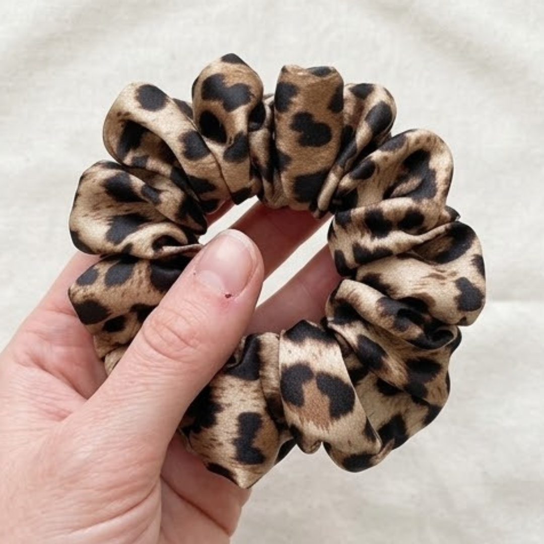 scrunchie-cetim-leopardo-detalhe-artesanal.jpg Close-up de mão segurando um scrunchie volumoso em cetim com padrão animal print de leopardo, destacando a textura acetinada e o acabamento artesanal sobre fundo neutro.