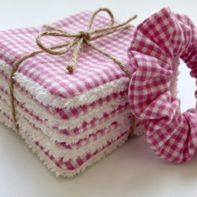 "Fotografia em grande plano de um conjunto de discos desmaquilhantes reutilizáveis e um scrunchie. Os discos são quadrados, feitos de tecido com padrão vichy rosa e branco de um lado, e um tecido atoalhado branco e volumoso do outro. Estão empilhados e atados com um laço de cordel de juta castanho. À direita, parte de um scrunchie com o mesmo padrão rosa está em foco, mostrando a textura do franzido do tecido. O fundo é branco e luminoso, realçando as cores vibrantes do conjunto."