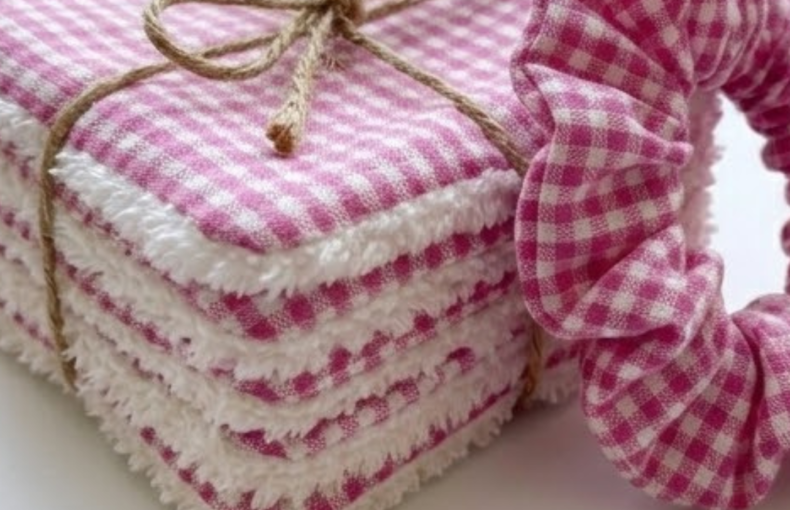 "Fotografia em grande plano de um conjunto de discos desmaquilhantes reutilizáveis e um scrunchie. Os discos são quadrados, feitos de tecido com padrão vichy rosa e branco de um lado, e um tecido atoalhado branco e volumoso do outro. Estão empilhados e atados com um laço de cordel de juta castanho. À direita, parte de um scrunchie com o mesmo padrão rosa está em foco, mostrando a textura do franzido do tecido. O fundo é branco e luminoso, realçando as cores vibrantes do conjunto."