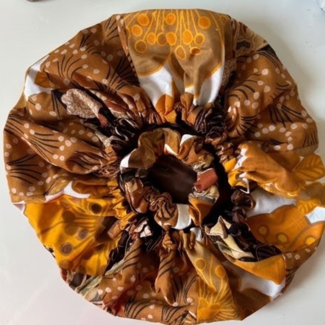 Vista superior de uma touca de dormir de cetim (bonnet) aberta, exibindo um padrão africano em tons de mostarda, castanho e branco com o interior em cetim castanho escuro.