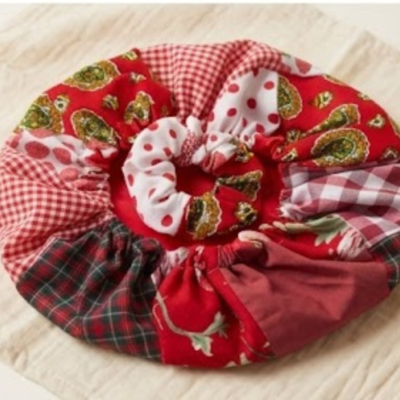 Vista superior de um conjunto capilar composto por uma touca de cetim (bonnet) e um elástico de cabelo (scrunchie) coordenado, ambos com padrão patchwork em tons de vermelho, branco e verde, sobre um tecido de linho claro.