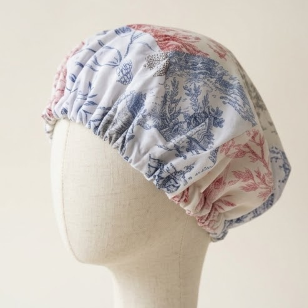 Close-up de uma touca de dormir de cetim (bonnet) com padrão Toile de Jouy em patchwork azul e vermelho coral, ajustada num manequim de busto de linho claro.