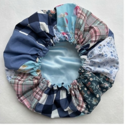 Uma touca de cetim feita à mão com técnica de patchwork em tons de azul, xadrez e floral, exibida de cima sobre um fundo branco.