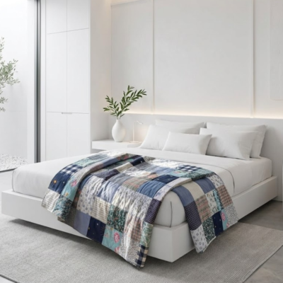 Uma cama de casal minimalista com lençóis brancos, destacando uma colcha de retalhos (patchwork) em tons de azul, marinho e cinzento disposta aos pés da cama.