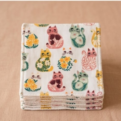 Fotografia de estúdio com fundo neutro bege claro, mostrando uma pilha perfeitamente organizada de toalhetes faciais reutilizáveis quadrados em tecido artesanal, destacando o padrão frontal de gatinhos ilustrados coloridos e o acabamento cuidado da costura branca nas bordas.