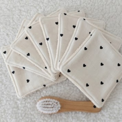 "Fotografia de estúdio com fundo texturado branco e luz natural suave, mostrando em detalhe um conjunto de toalhetes e discos desmaquilhantes reutilizáveis dispostos em leque, conforme a imagem Cópia de ETSY produto (44).png. O tecido é de algodão branco natural com padrão minimalista de pequenos corações pretos. À frente dos discos, encontra-se uma pequena escova de limpeza facial com cabo de madeira clara."
