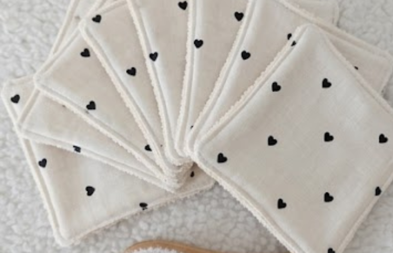 "Fotografia de estúdio com fundo texturado branco e luz natural suave, mostrando em detalhe um conjunto de toalhetes e discos desmaquilhantes reutilizáveis dispostos em leque, conforme a imagem Cópia de ETSY produto (44).png. O tecido é de algodão branco natural com padrão minimalista de pequenos corações pretos. À frente dos discos, encontra-se uma pequena escova de limpeza facial com cabo de madeira clara."