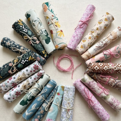 Rolos de retalhos de tecidos de algodão com padrões florais, botânicos e geométricos de estilo vintage e cottagecore, organizados em círculo perfeito sobre uma mesa de linho claro e rústico. No centro do círculo, destaca-se um pequeno novelo de fio rosa.